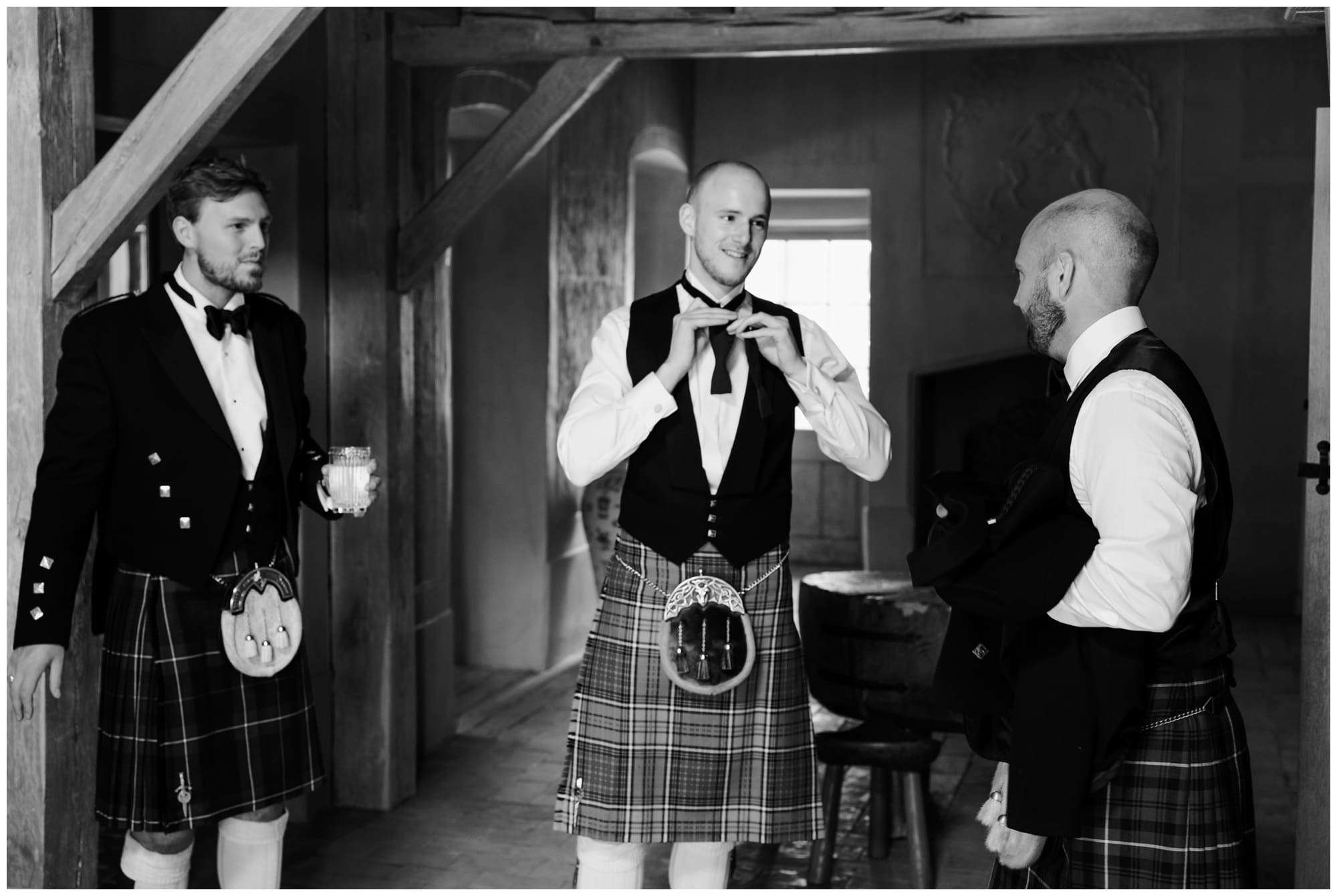 Groom in kilt doing his bowtie up with his groomsman.